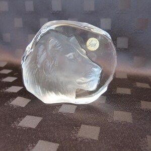 May include: A clear, sculpted glass lion head paperweight. The lion's profile is detailed, with a textured mane and facial features. A small, round sticker is affixed to the glass. The paperweight is a decorative item.