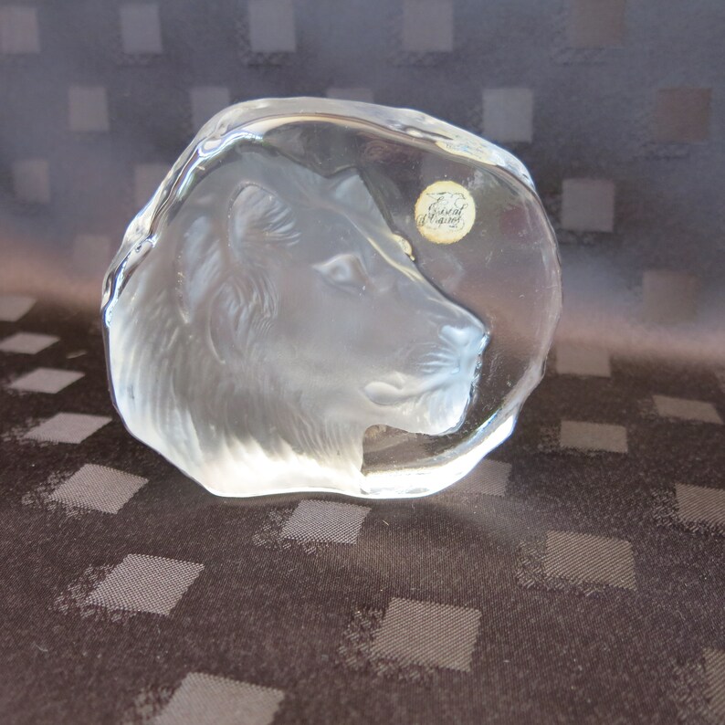 May include: A clear, sculpted glass lion head paperweight. The lion's profile is detailed, with a textured mane and facial features. A small, round sticker is affixed to the glass. The paperweight is a decorative item.