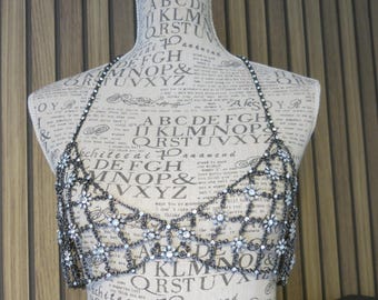 Op de afbeelding: Een decoratieve, donkere metalen lichaamsketting met een bloemen- en geometrisch ontwerp. De ketting is versierd met kleine, heldere kristallen. Het stuk wordt getoond op een mannequin met bedrukte tekst.