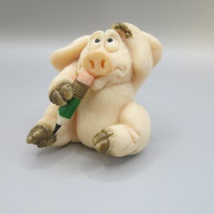 Könnte beinhalten: Eine kleine, weiße Keramikfigur eines Schweins, das einen grün-weißen Bleistift hält. Das Schwein hat eine rosa Nase und große, braune Augen. Das Schwein sitzt mit erhobenem rechten Bein und hält den Bleistift in der linken Hand.