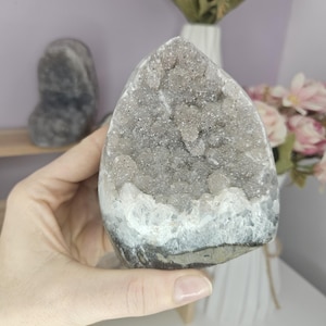 May include: A grey geode crystal with a sparkling interior and a white base. The crystal is held in a hand, with a blurred background featuring flowers and another geode.