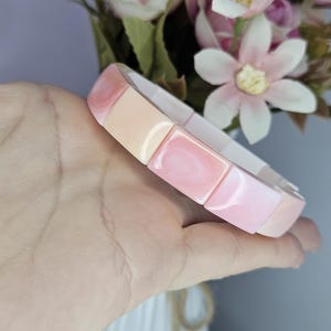 May include: A stretch bracelet composed of rectangular, smooth, glossy segments in alternating shades of soft pastel pink, peach, and cream. The individual pieces are linked together to form a continuous band with a polished, reflective finish.