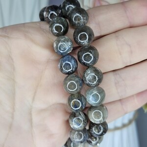 May include: Two beaded bracelets featuring iridescent, grey-green, and blue spherical beads. The beads have a reflective, circular pattern. The bracelets are held in a hand, with a blurred floral background.