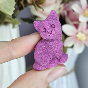 May include: A small, textured, vibrant purple cat-shaped object. The figure features a seated feline silhouette with simple, indented circular eyes and a small cross-shaped mark representing the nose and mouth area, crafted with a granular surface.