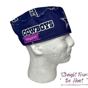 May include: A navy blue scrub cap featuring the word "COWBOYS" in white block letters and a white star. The cap also has the text "Straight From the Heart STUDIOS LLC" on a purple tag.