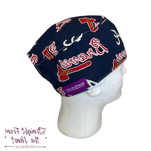 May include: A navy blue cap featuring the Atlanta Braves logo in red and white. The cap has a repeating pattern of the team's logo and wordmark. A small purple tag with text is attached to the cap.