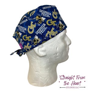May include: A navy blue scrub cap featuring a repeating pattern of gold and white Georgia Tech logos and yellow jackets. The cap is tied at the back and is displayed on a white mannequin head.