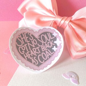 May include: A pink heart-shaped sticker with a white border and white stars. The sticker has black text that reads "She's got no soul heart black as coal."