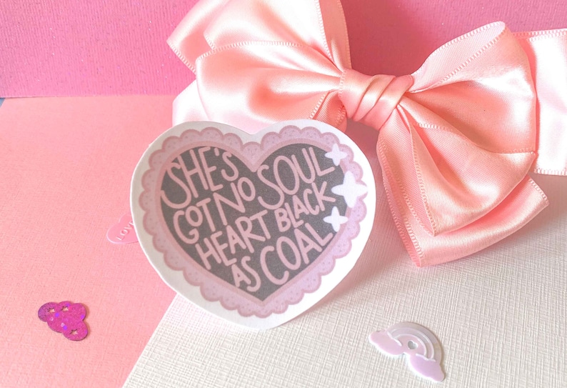 May include: A pink heart-shaped sticker with a white border and white stars. The sticker has black text that reads "She's got no soul heart black as coal."