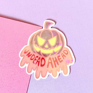 May include: A holographic sticker with a dripping orange pumpkin and the text "UNDEAD AHEAD" in red.