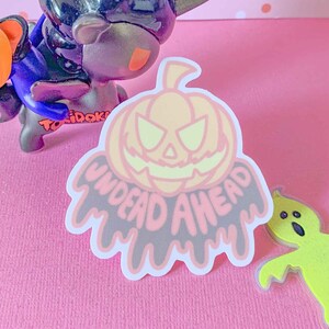 May include: A pink and white sticker with a cartoon pumpkin face and the text "UNDEAD AHEAD" in dripping black letters.