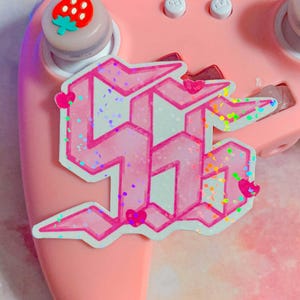 May include: A pink and white sticker with a glittery finish, featuring a stylized number "56" in a bold, geometric font. The sticker is attached to a pink gaming controller.