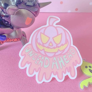 May include: A pink and white sticker with a dripping, melting pumpkin design. The pumpkin has a scary face with yellow eyes and a wide, toothy grin. The text "UNDEAD AHEAD" is written in a dripping, melting font below the pumpkin.
