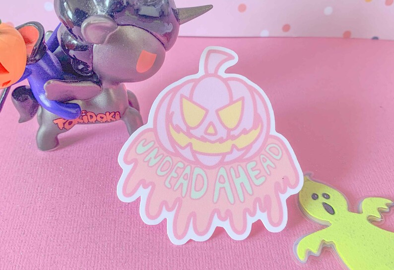 May include: A pink and white sticker with a dripping, melting pumpkin design. The pumpkin has a scary face with yellow eyes and a wide, toothy grin. The text "UNDEAD AHEAD" is written in a dripping, melting font below the pumpkin.