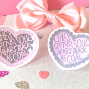 May include: Two pink and gray heart-shaped stickers with the text "She's got no soul heart black as coal" on each. The stickers are on a pink and white surface.