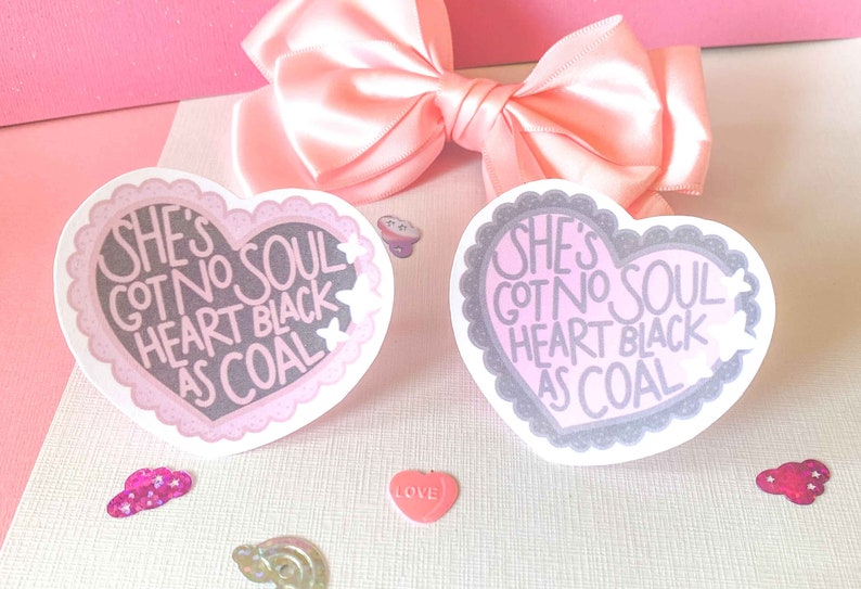 May include: Two pink and gray heart-shaped stickers with the text "She's got no soul heart black as coal" on each. The stickers are on a pink and white surface.