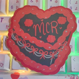 May include: A red and black heart-shaped sticker with the letters "MCR" in white inside. The sticker has a cake-like design with white and gray bows around the edges.
