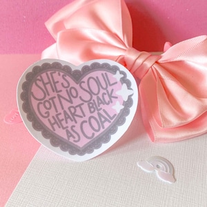 May include: Pink heart-shaped sticker with the text "She's got no soul heart black as coal" in white lettering. The sticker has a gray border and is decorated with white stars.