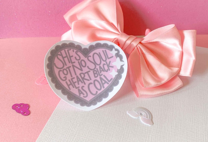 May include: Pink heart-shaped sticker with the text "She's got no soul heart black as coal" in white lettering. The sticker has a gray border and is decorated with white stars.