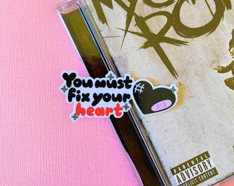May include: A black and white sticker with the text "You must fix your heart" and a black heart with a pink bandage on it. The sticker is on a CD case with a parental advisory label.
