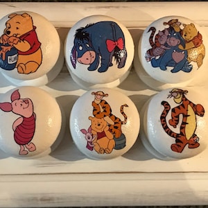 May include: Six white round knobs with different Winnie the Pooh characters printed on them. The characters include Winnie the Pooh, Piglet, Tigger, Eeyore, and a group of characters including Tigger, Piglet, and Winnie the Pooh.