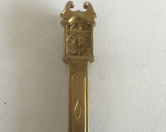 May include: A gold-colored, miniature grandfather clock with a Roman numeral clock face. The clock has a diamond-shaped design on the front.