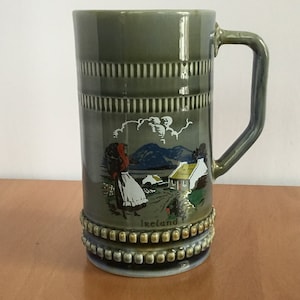 Puede incluir: Una taza de cerámica verde con una escena de una mujer caminando en un paisaje rural irlandés. La taza tiene un asa y un borde de cuentas. Las palabras "Irlanda" están impresas en la parte inferior de la taza.