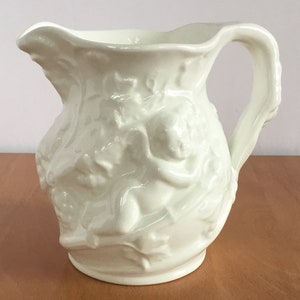 May include: A white ceramic pitcher with an embossed design of a cherub and floral motifs. The pitcher has a wide mouth and a handle.