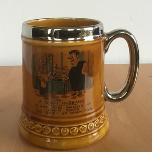 May include: A brown ceramic mug with a silver rim and handle. It features a cartoon illustration of a man washing dishes with the text "A good husband is one who washes up when asked and dries up when told."