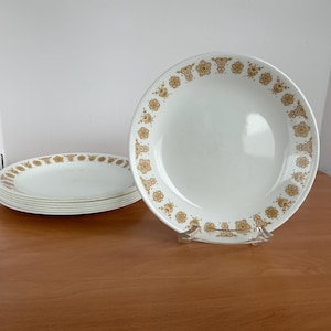 以下が含まれることがあります： 縁に金色の花柄模様が入った白いセラミックプレートのセット。 プレートは積み重ねられ、木製の表面に表示されています。 食事を提供するクラシックなデザインです。