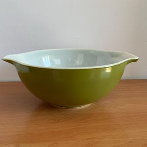 Op de afbeelding: Een ronde mengkom met een gladde olijfgroene buitenkant en een glanzende witte binnenkant. De kom is voorzien van twee geïntegreerde schenktuiten aan weerszijden, wat zorgt voor een breed, ondiep silhouet met een gebogen basis en een strakke afwerking.
