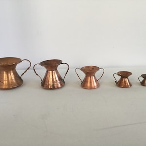 Puede incluir: Conjunto de cinco tazas medidoras de cobre con dos asas. Las tazas están graduadas en tamaño, siendo la taza más grande la más alta y la taza más pequeña la más baja.