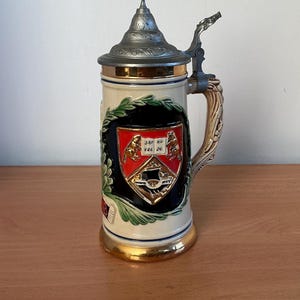 May include: A ceramic beer stein featuring a metallic pewter lid with a thumb lever. The body is cream-coloured with a central crest displaying two animals flanking an open book with the text SAP AU ERE DE, set against a dark blue background.