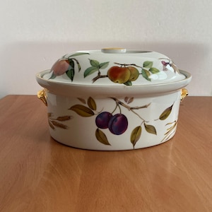 May include: A white ceramic casserole dish with a lid, adorned with a fruit design featuring plums, apples, and peaches. The dish has gold-colored handles and a matching gold accent on the lid's knob. A decorative kitchenware item.