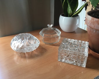 May include: Three clear glass decorative boxes with intricate cut designs. The boxes are on a wooden surface.