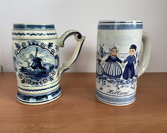 May include: Two ceramic beer steins with blue and white designs. The left stein features a windmill in a decorative frame. The right stein depicts children in traditional clothing with a windmill in the background and the word Holland printed below.