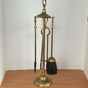 May include: A brass fireplace tool set with a stand. The set includes a poker, tongs, a small broom, and a shovel. The tools are suspended from a central stand with a circular base. The stand is approximately 76 cm tall.