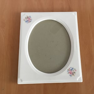Puede incluir: Marco de fotos de cerámica blanca con una abertura ovalada y diseños florales. El marco tiene un fondo blanco con flores rosas, azules y verdes.