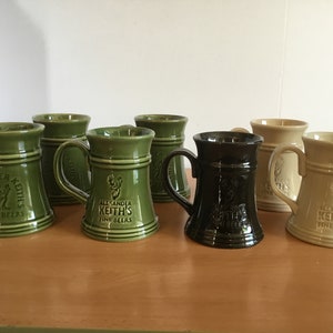 May include: A selection of ceramic beer tankards in shades of green, black, and beige. Each tankard has a handle and the text "Alexander Keith's Fine Beers". The tankards are displayed on a wooden surface.