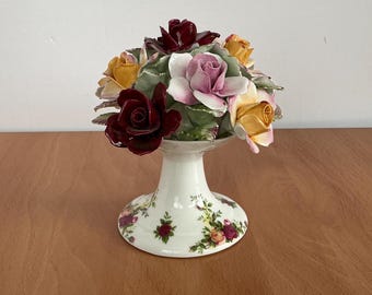 May include: A white ceramic pedestal with a floral pattern holds a bouquet of porcelain roses. The roses are in shades of pink, yellow, and burgundy.