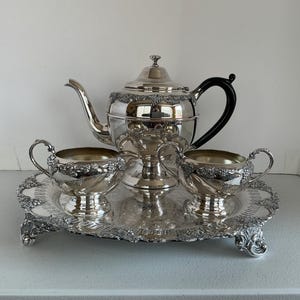 May include: A silver-plated tea set comprising a teapot with a black handle, a sugar bowl, and a creamer, all featuring embossed grape and vine motifs. The set is arranged on a matching silver-plated serving tray with ornate feet and edges.