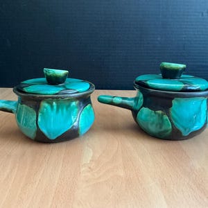 May include: Two ceramic lidded pots with handles. The pots are teal and brown with a floral design. The lids have a small knob on top. The pots are sitting on a wooden surface.
