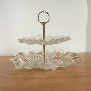 May include: Two-tiered glass serving stand with a silver metal base and a ring handle. The glass plates have a scalloped edge.