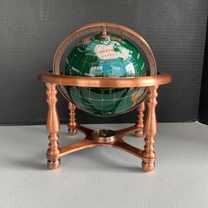May include: A decorative globe featuring a deep green ocean surface with inlaid continents in various stone colours. The globe is mounted within a copper-toned metal frame with a circular meridian ring and four turned legs, including a small compass.