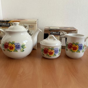 Könnte beinhalten: Ein weißes Keramik-Teeservice mit Teekanne, Zuckerdose und Sahnekännchen, alle mit einem floralen Stiefmütterchen-Design in Rot-, Gelb- und Lilatönen verziert. Das Set steht auf einer Holzoberfläche.
