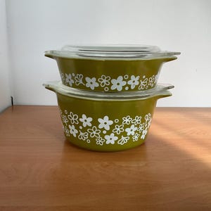 May include: Two stacked olive green glass casserole dishes with integrated side handles and clear glass lids. The exterior of each bowl features a repeating white floral pattern consisting of five-petaled flowers and small circular dots.