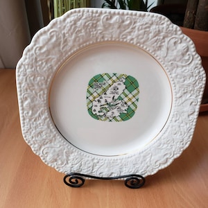 Puede incluir: Plato de cerámica blanco con borde dorado y un diseño floral en relieve. El centro del plato presenta un mapa de la isla de Cabo Bretón, Nueva Escocia, Canadá, con un fondo de tartán verde y amarillo.