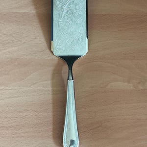 Peut inclure: Une pelle à gâteau argentée avec une lame rectangulaire ornée d'un motif floral en relief. Le manche est long et se termine par une extrémité arrondie. Posée sur une surface en bois.