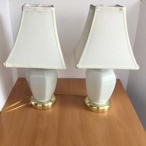 May include: A pair of white ceramic table lamps with white fabric lampshades. The lamps have gold bases and are sitting on a wooden surface.