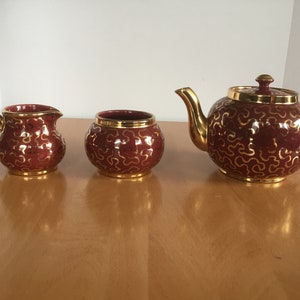 May include: A red tea set with gold accents. The set includes a teapot, creamer, and sugar bowl. The teapot has a round body and a spout on the side. The creamer and sugar bowl are both round.